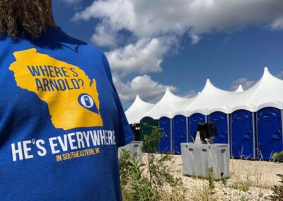 An Arnold's team member wearing an awesome #Where'sArnold t-shirt in front of our Weekender Portable Restrooms and 2-Sided Hand Wash Stations.